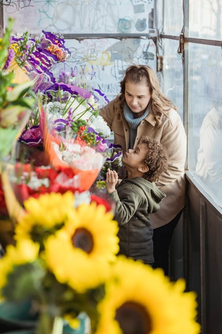 A woman and a young girl are shopping for fresh flowers in a bright, greenhouse-style floral shop. The woman has light brown, curly hair and is wearing a beige coat over a blue scarf, gently smiling a