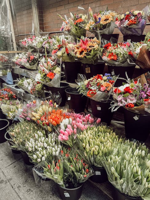 A display of numerous flower bouquets arranged on black shelves and large buckets outdoors, featuring a variety of fresh flowers in vibrant colors such as pink, orange, red, purple, and white. The flo