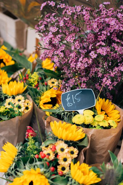 A vibrant assortment of fresh flower bouquets displayed outdoors at a flower market. The arrangements include bright yellow sunflowers, small white daisies, and red blossoms, all wrapped in brown craf