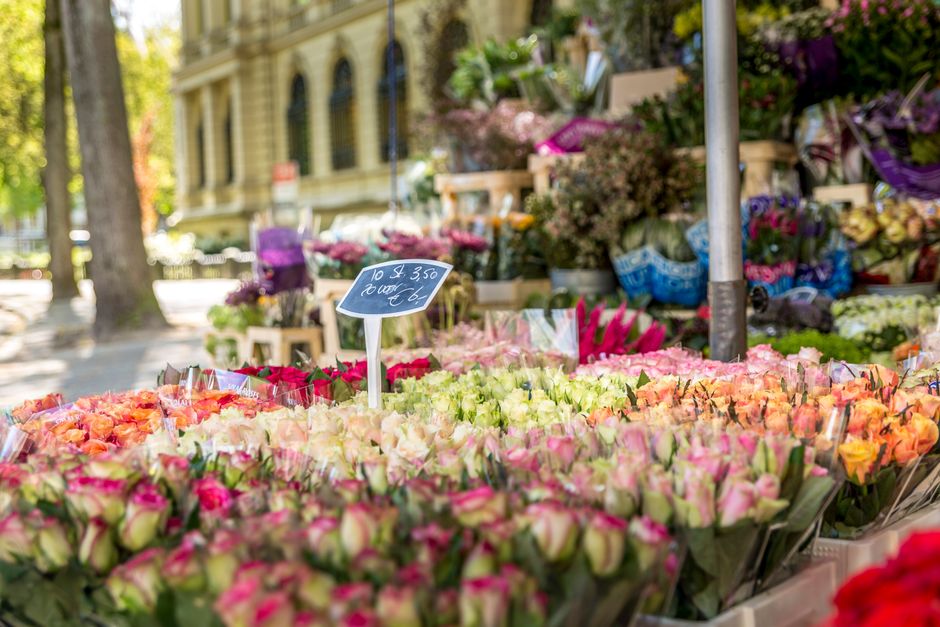 A vibrant outdoor market stall displaying an extensive array of fresh flower bouquets and floral arrangements. In the foreground, numerous tightly packed Roses in shades of pink, peach, and cream are 