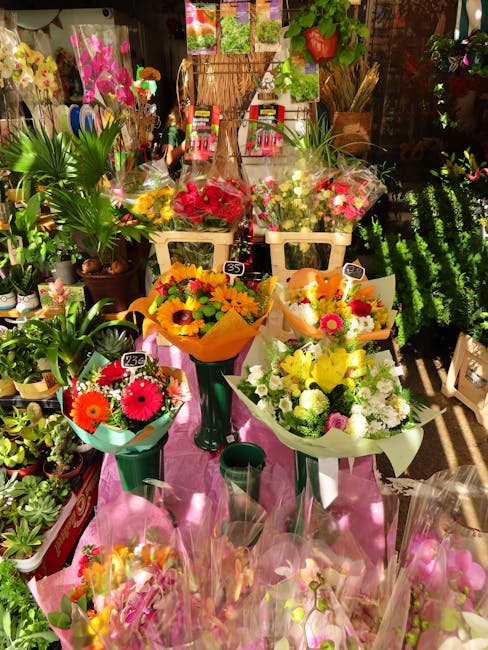 A display of fresh flower bouquets arranged on tall green stands and a pink table inside a florist shop. The bouquets feature bright, vibrant flowers including sunflowers, pink gerbera daisies, white 