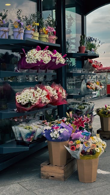 A display of fresh floral arrangements and bouquets situated outside a florist shop on a multi-tiered shelving unit. The arrangements include white, pink, and purple flowers, with some wrapped in pink