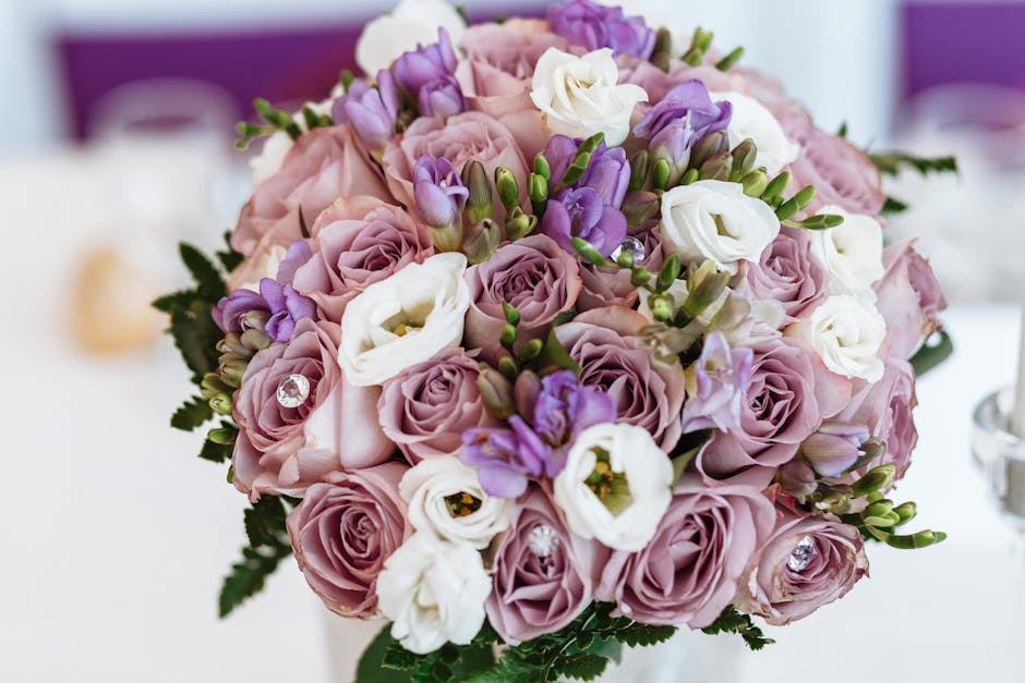 A close-up of a floral gift featuring a bouquet with lavender pink roses, white lisianthus, and purple freesias, arranged with lush green foliage. The bouquet is densely packed, with the flowers arran